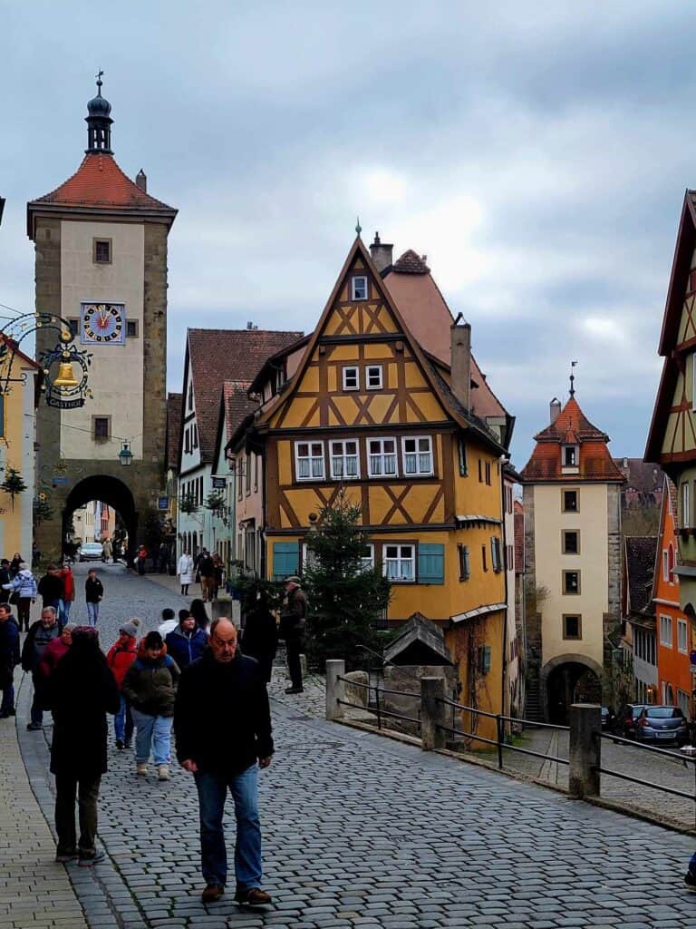 A forked cobblestone street with a gated tower on the left, an orange medieval timbered house in the center, and another medieval building on the right. Lots of people milling around