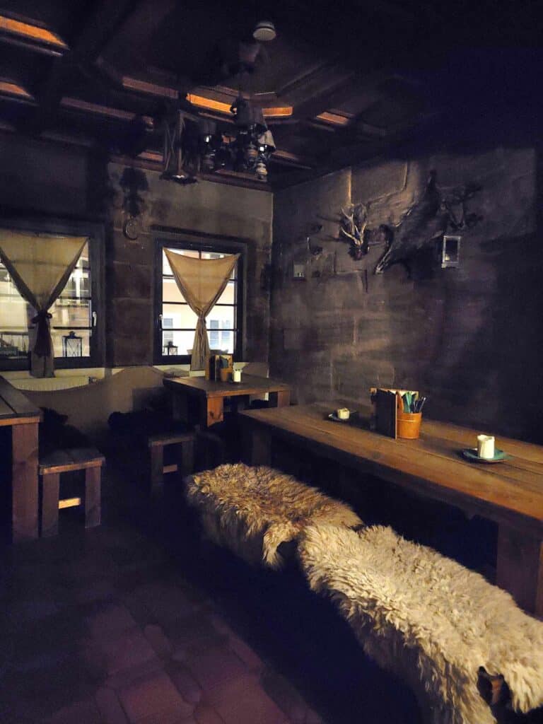 A dimly lit stone room with long wooden benches with candles on the tables and sheepskin to sit on