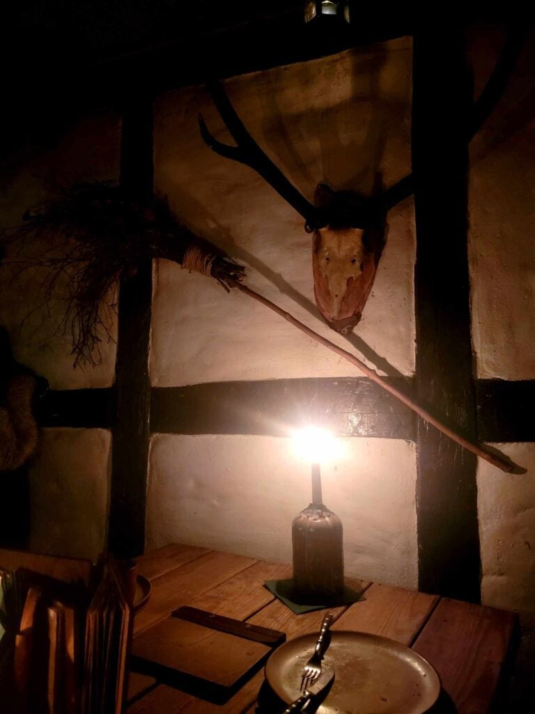 A candle lights a place setting with a pewter serving dish, twisted silverware, and a timbered wall on which hangs a deer skull and antlers