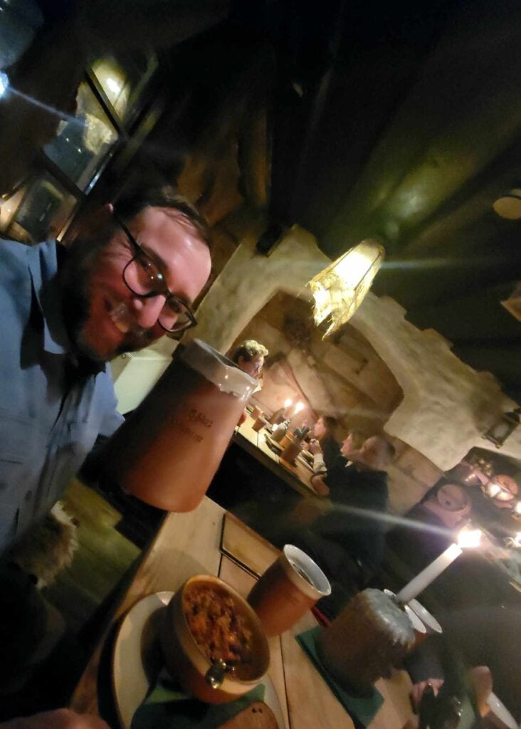 A smiling man with a gigantic earthenware tankard of beer, and a bowl of stew