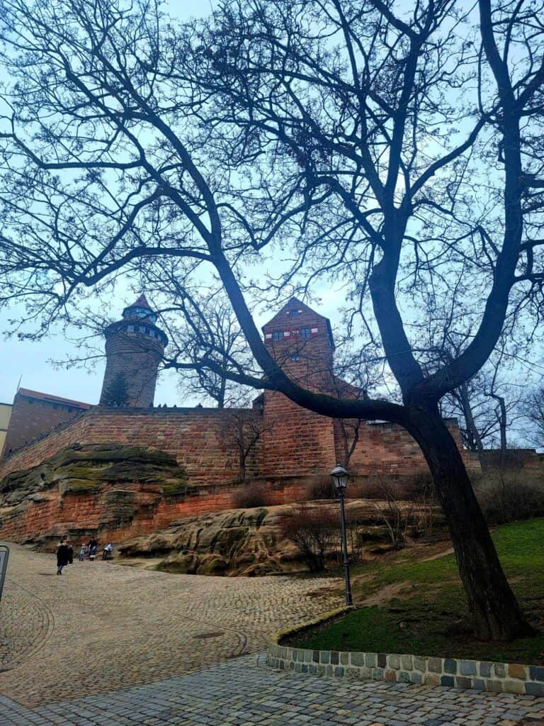 A high sandstone wall with two towers at the top of a hill with a cobblestone street leading to it and a large tree in winter in the foreground