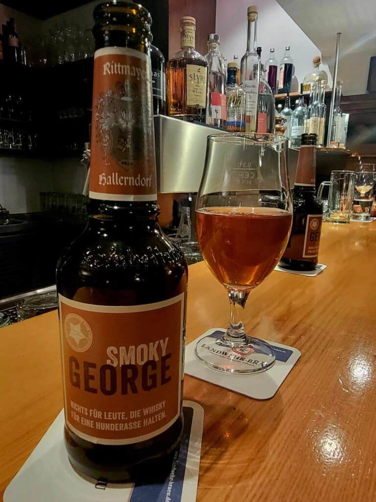 A bottle of beer labeled Smoky George and a beer glass half full of amber beer sit on a wooden bar