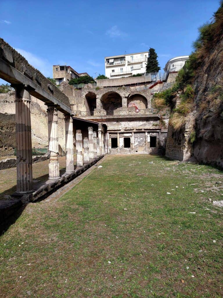 Pompeii vs. Herculaneum: Best Tips & Biggest Differences 7 A long rectagonal space with dirt and short grass flanked by a row of columns