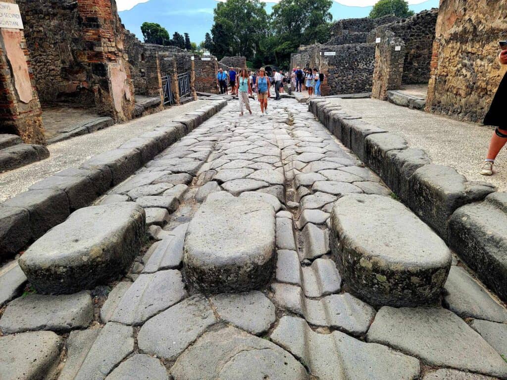 Pompeii vs. Herculaneum: Best Tips & Biggest Differences 14 A stone road in Pompeii showing grooves for chariot wheels, gutters for water runoff, curbs, and large stones to cross the street