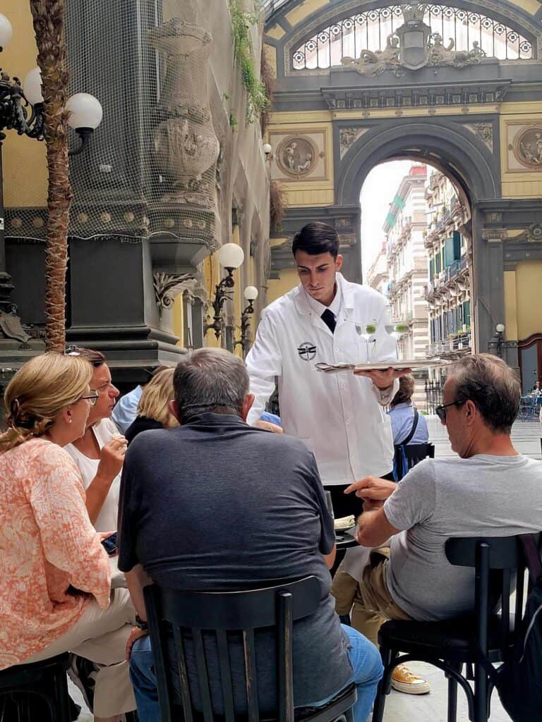 Pompeii vs. Herculaneum: Best Tips & Biggest Differences 29 A dark-haired man in a white coat and black tie carries a tray of martinis with olives which he serves to a table of four inside a gorgeous covered market hall in Naples