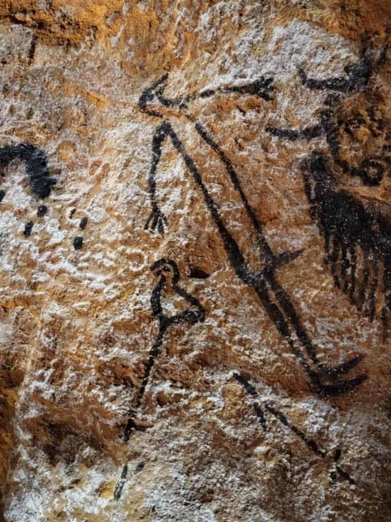 Best 2 Day Dordogne Prehistory Itinerary: What to See & Do 16 A dark rudimentary charcoal outline of a human figure with arms outstretched and a bird head. It stands between a bird on a stick and an ourocs