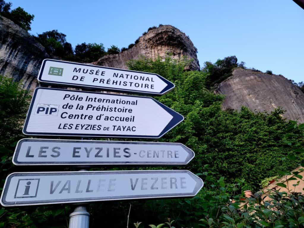 Best 2 Day Dordogne Prehistory Itinerary: What to See & Do 2 A set of stacked directional street signs reading: Musee National de Prehistoire; Pole International de la Prehistoire Centre d'accueil; Les Eyzies - Centre; and Vallee Vezere, with a dramatic cliff behind