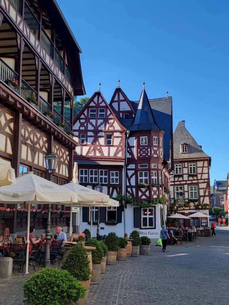 One Day in Bacharach: Best Guide to Germany's Hidden Gem on the Rhine 19 A series of half timbered buildings on a cobblestone street, tables with diners underneath umbrellas, and potted plants line the road